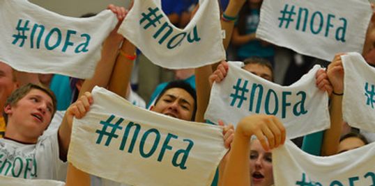 Energetic students hold up white cloth banners with '#NOFA' written on them in blue text at the 5th annual NOFA Game